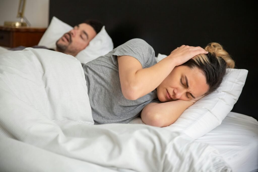 A woman looks frustrated in bed as her partner snores loudly, highlighting sleep disturbance issues.